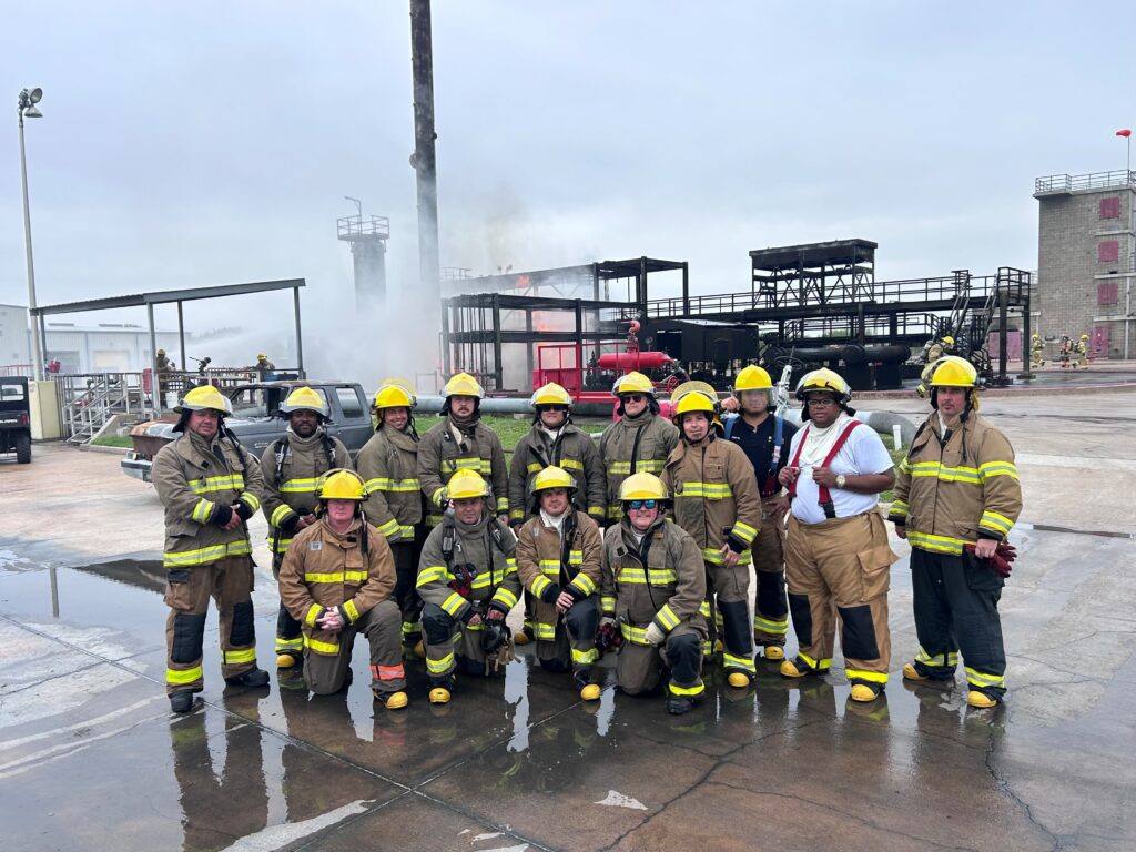 an emergency response team. 14 men in fireproof suits with safety gear standing in front of a fire fighting training facility