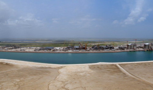 Rio Grande LNG construction site with cranes and storage tanks across a turquoise ship channel, coastal flats beyond.